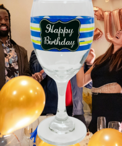 Happy Birthday Striped Wine Glass displayed against a lively birthday party with smiling guests, gold balloons, and purple flowers.