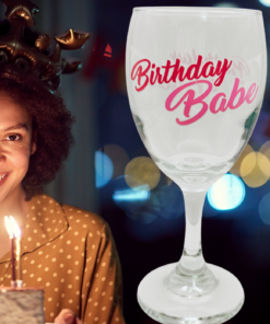 Birthday Babe Pink Glitter Wine Glass displayed beside a smiling woman wearing a birthday crown and holding a candlelit birthday cake.