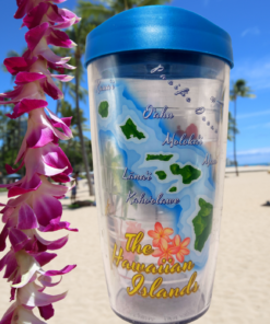 Hawaiian Islands Map Blue Lid Travel Tumbler displayed beside purple tropical flowers against a sunny Hawaiian beach with palm trees.