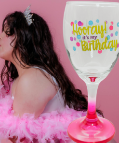Hooray It's My Birthday Pink Stem Wine Glass displayed beside a birthday woman wearing a tiara and holding a pink strawberry cake.