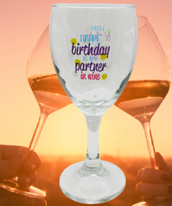 Happy Birthday Partner In Wine Glass displayed against a golden sunset with two people raising wine glasses in a celebratory toast.