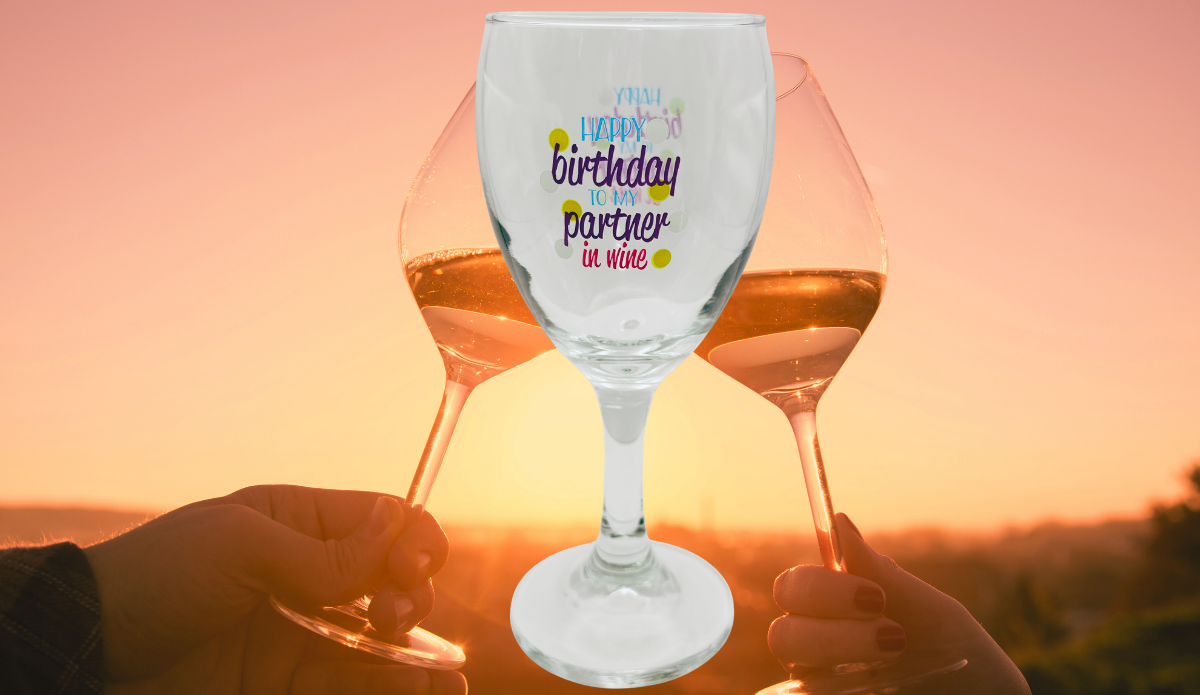 Happy Birthday Partner In Wine Glass displayed against a golden sunset with two people raising wine glasses in a celebratory toast.