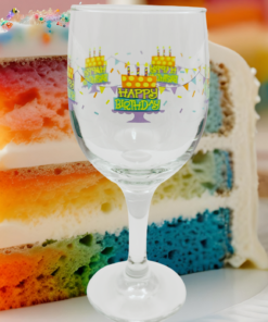 Happy Birthday Cake Wine Glass displayed in front of a colorful slice of layered birthday cake with pink, orange, yellow, and blue cake layers.