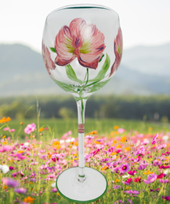 Hand Painted Dark Pink Flower Balloon Wine Glass displayed in a colorful meadow of pink, orange, and white wildflowers with green hills and distant mountains.