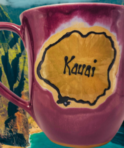 Kauai Burgundy Souvenir Mug displayed against Kauai Nāpali Coast mountains and turquoise ocean with golden island silhouette.