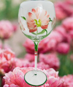 Hand Painted White Pink Flower Balloon Wine Glass displayed among soft pink flowers, featuring creamy white petals with coral-pink accents, green leaves, and a green-edged foot.