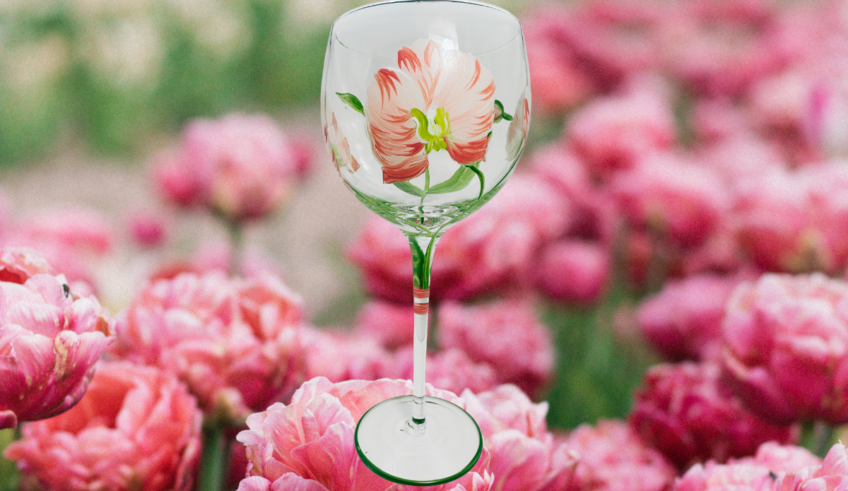 Hand Painted White Pink Flower Balloon Wine Glass displayed among soft pink flowers, featuring creamy white petals with coral-pink accents, green leaves, and a green-edged foot.